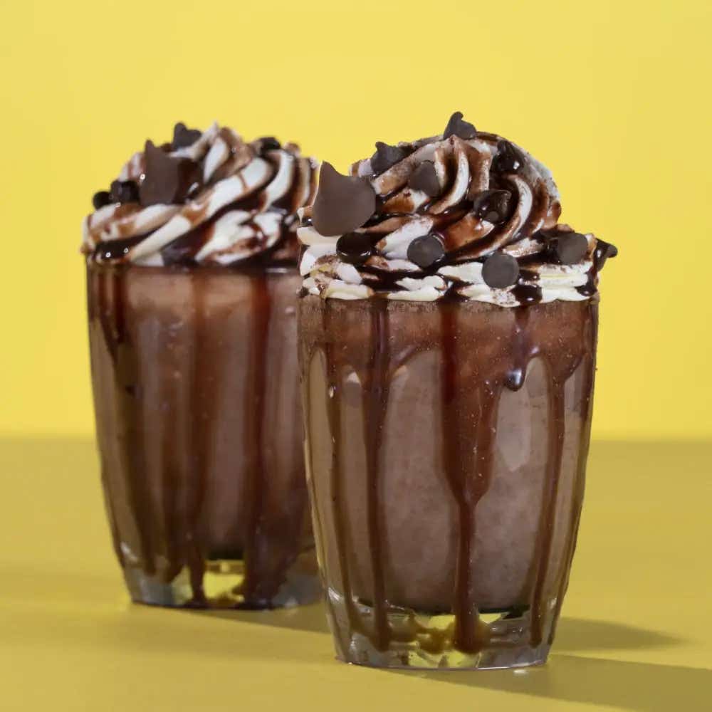 Dos vasos de cristal llenos de frappe decorados con crema batida, KISSES de Leche y Jarabe de Chocolate HERSHEY'S.