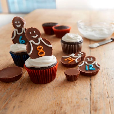 Cupcakes pequeños sobre una mesa de madera, decorados con crema batida y REESE'S formando y decorados para que parezcan jugadores de fútbol.