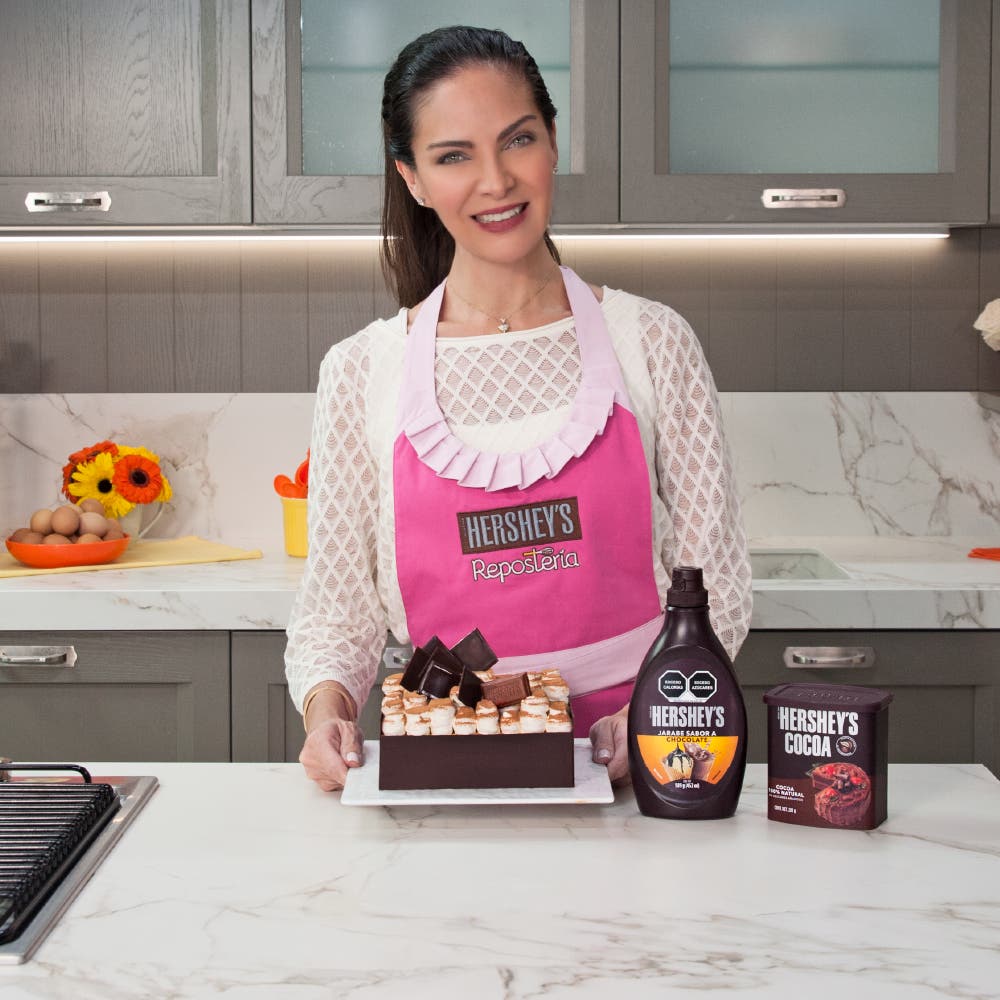 Paulina Abascal sonriendo en una cocina mientras usa un delantal con el logo de HERSHEY’S. Frente a ella, en la mesa, se encuentran varios productos HERSHEY’S para repostería.