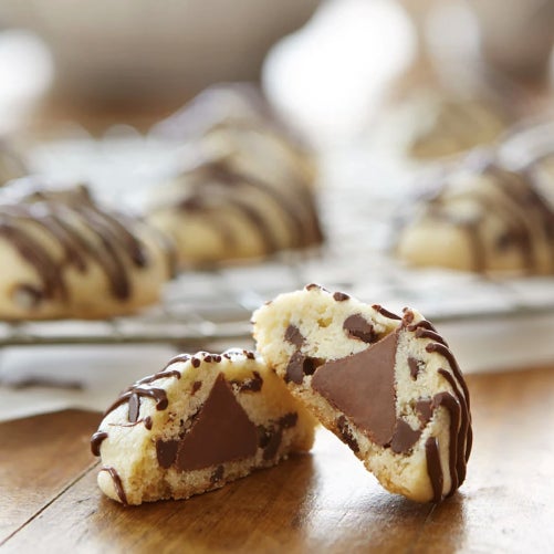 Se ve una tabla de madera con una galleta con líneas de chocolate partida por la mitad, en el corazón de la galleta deja ver un Kiss dividido por la mitad que se encuentra dentro, al fondo se ve un plato con más galletas sin partir.