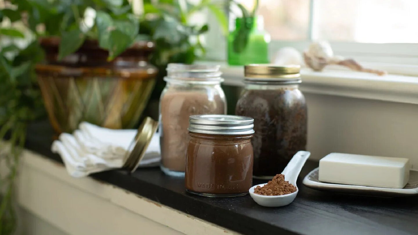 Se ven 3 frascos con productos para spa con base de chocolate HERSHEY'S, una cuchara blanca con polvo de chocolate, una barra de jabón, unas toallitas blancas y unas plantas al fondo.