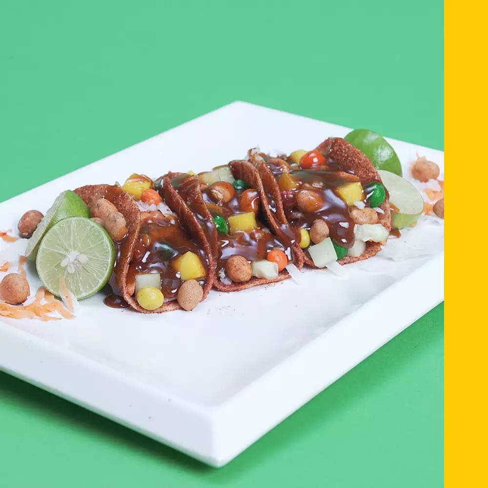 Tacos de pepino enchilado rellenos de jícama, zanahoria, pelón pelontes y cacahuates joponeses, acompañados con medios limones sobre un plato plano ractangular blanco.