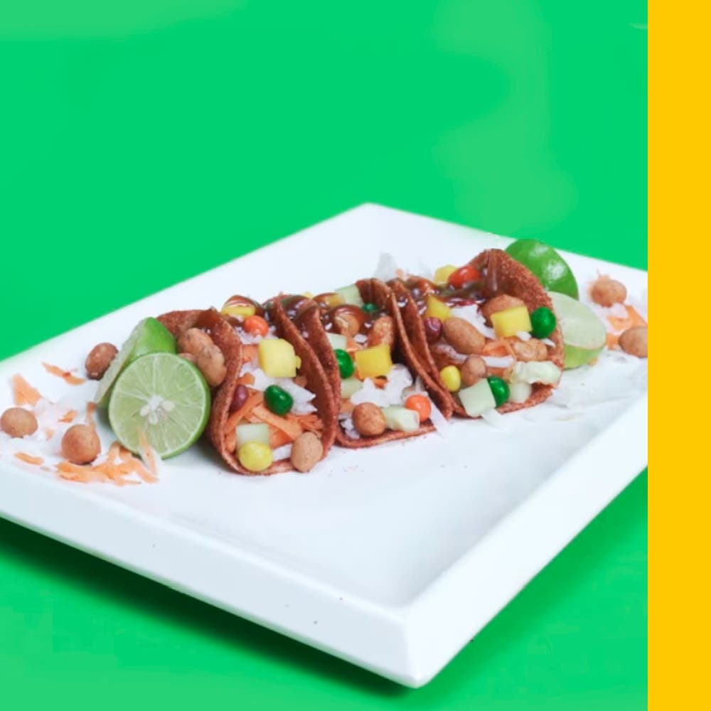 Tacos de pepino enchilado rellenos de jícama, zanahoria, pelón pelontes y cacahuates joponeses, acompañados con medios limones sobre un plato plano ractangular blanco.