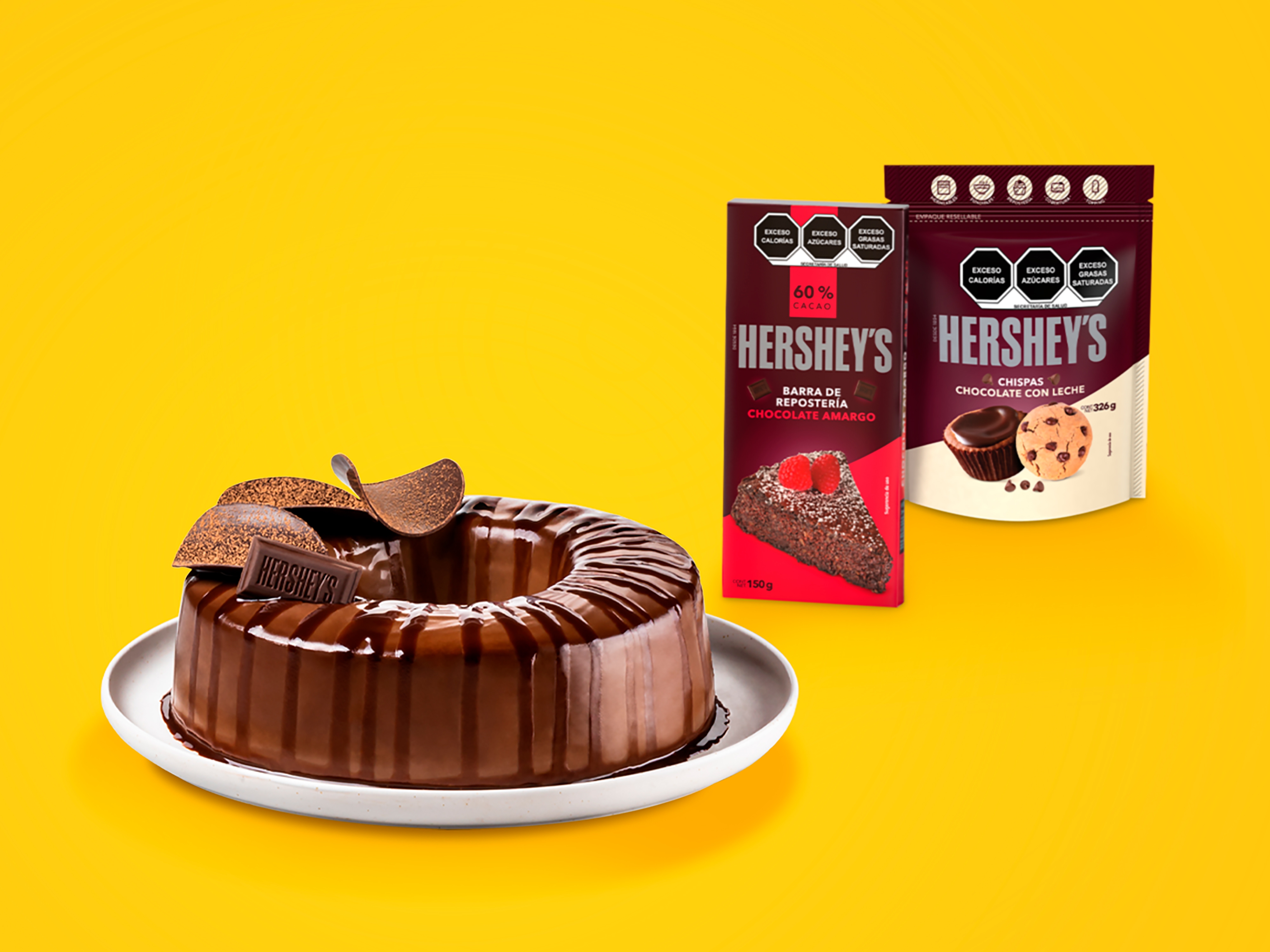 Plato blanco con una gelatina en forma de rosca de chocolate, bañada con chocolate líquido y adornada con Hershey's barra y láminas de chocolate. Fondo color amarillo y una barra de Repostería Hershey's Chocolate Amargo de 150 g y un empaque de chispas de chocolate con leche de 326 g