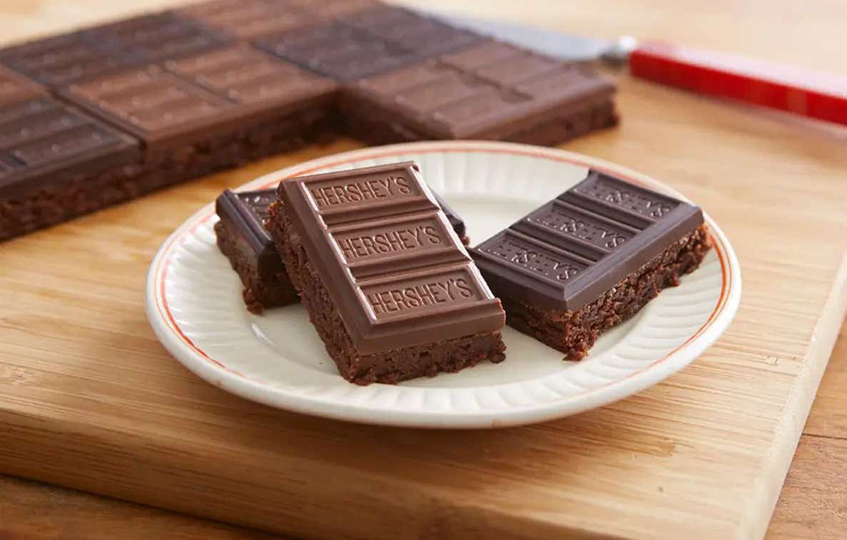 Sobre un base de madera, un plato color blanco con 3 trozos de brownie decorados con Barras de HERSHEY'S. Al fondo el resto de los brownies. 