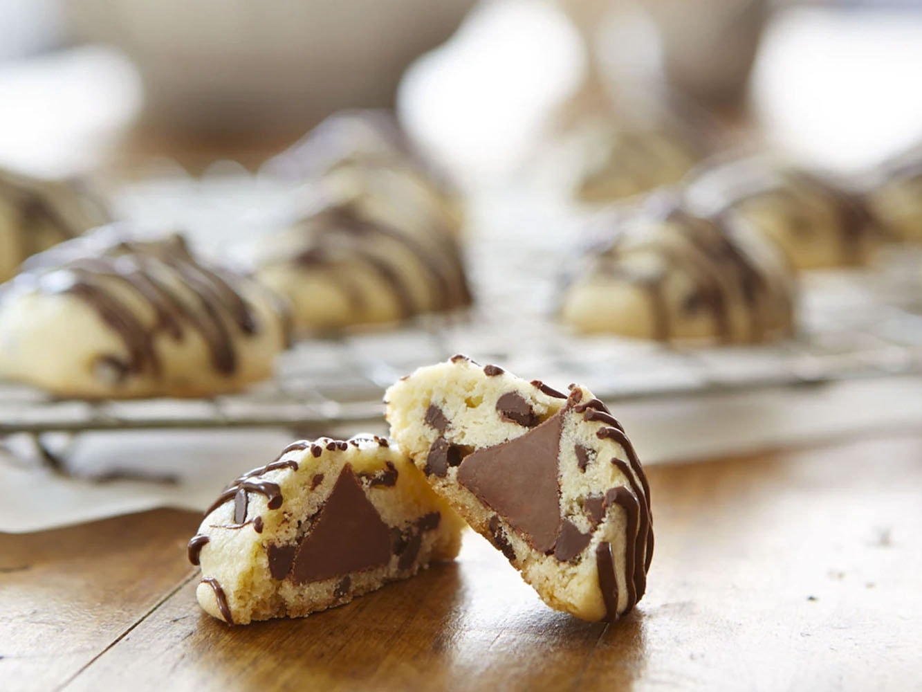 "Mesa de madera con una galleta partida a la mitad con un chocolate KISSES al centro, decoradas con rayas de chocolate. Al fondo, una charola con las mismas galletas.   "