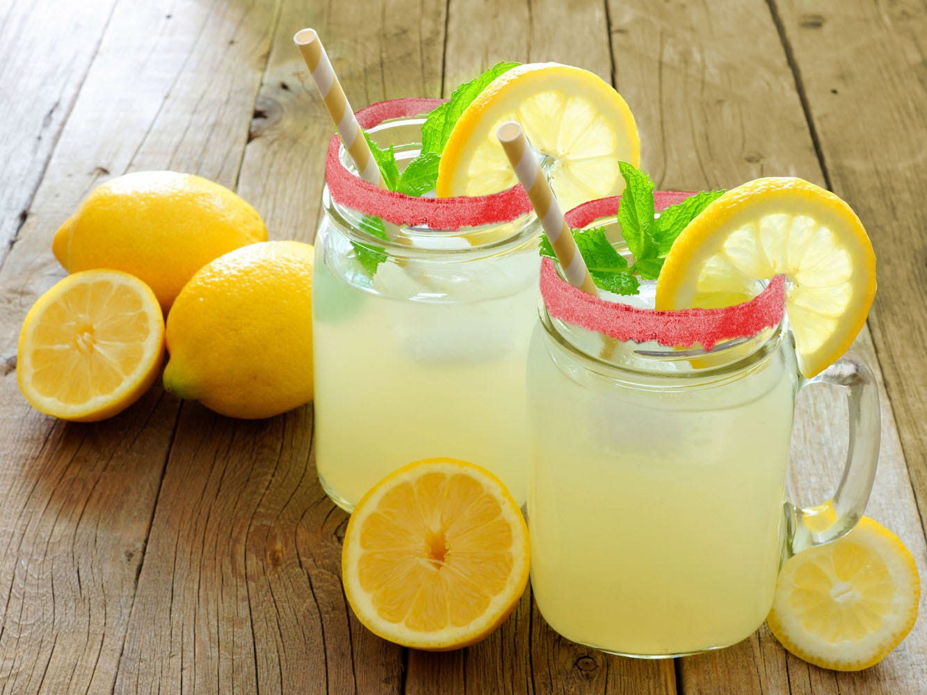 Sobre una mesa de madera, 2 frascos de cristal con limonada en el interior. Cada frasco decorado con una rodaja de limón, hojas de hierba buena y escarcha de crayón fresa. Alrededor de los frascos 2 limones enteros y 3 mitades. 
