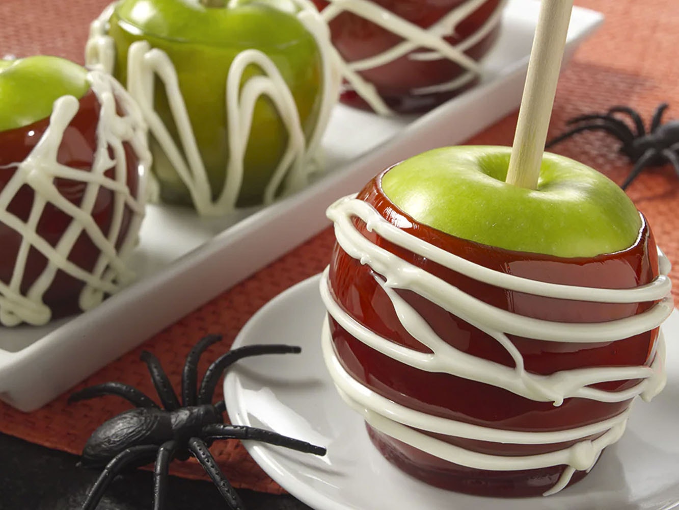 Sobre un mantel color rojo, un plato color blanco con una manzana verde forrada de caramelo color rojo y decorado de rayas blancas. Al fondo otro plato blanco con una manzana de caramelo rojo y una de caramelo verde. 