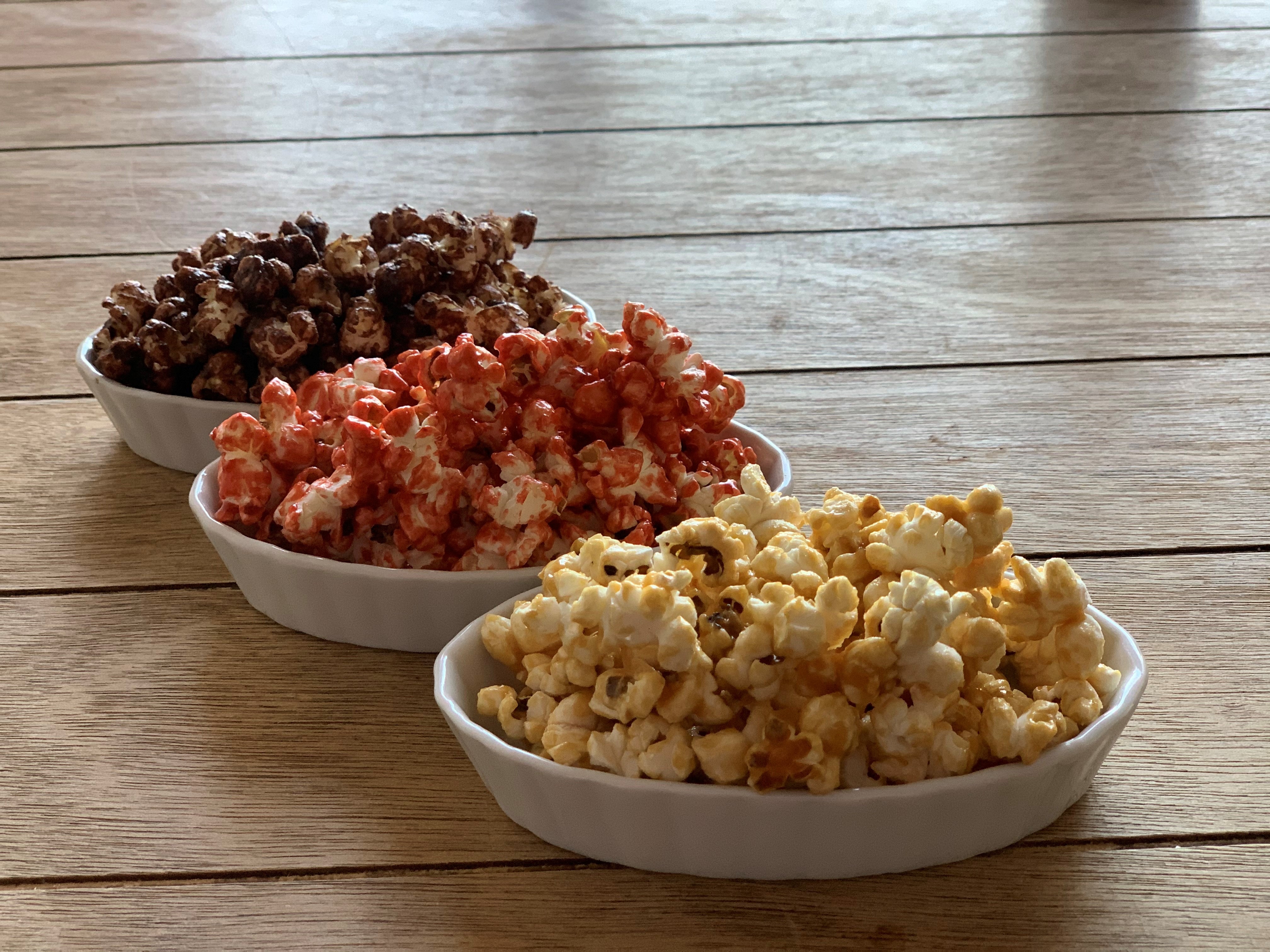 Sobre una mesa de madera 3 tazones. En el fondo un tazón con palomitas de maíz y chocolate, un tazón con palomitas de maíz y chile y al frente un tazón con palomitas de maíz y mantequilla. 