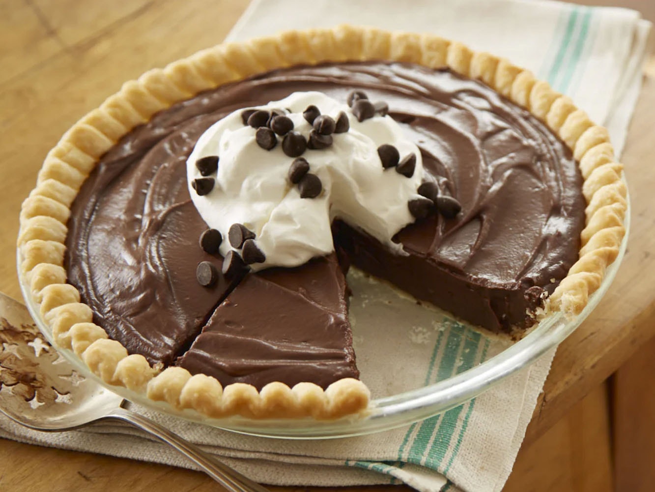 "Sobre una mesa de madera, un plato de cristal con un pay de chocolate, decorado con crema batida y chispas de chocolate.  "