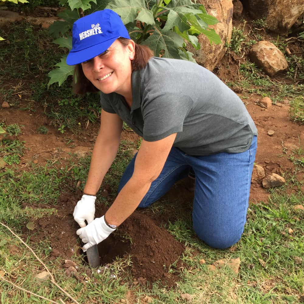 Mujer adulta usando una gorra con la marca Hershey's bordada, arrodillada en el pasto sembrando un árbol.