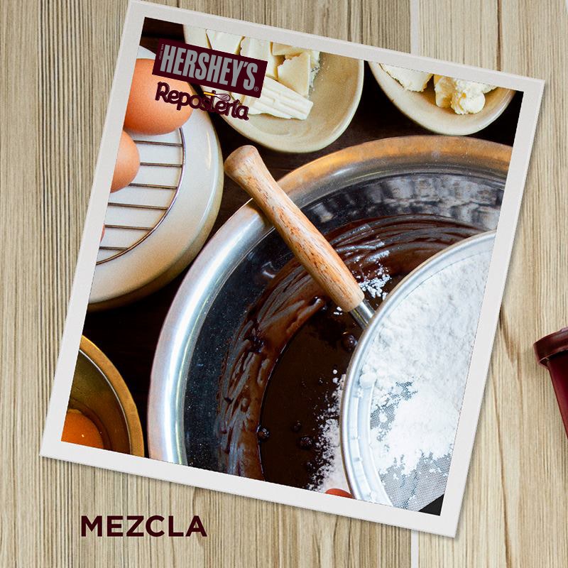 Mezcla. Sobre un fondo con textura de madera se ve una fotografía de un bowl metálico y un batidor lleno de Chocolate derretido. Al frente del bowl se alcanza a apreciar una Barra de Chocolate.