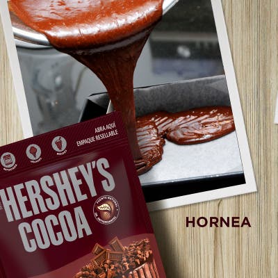 Hornea.  Sobre un fondo con textura de madera se ve una fotografía de un bowl metálico vaciando una mezcla de chocolate dentro de un recipiente para hornear. Frente a la fotografía se encuentra un empaque de Cocoa HERSHEY'S.