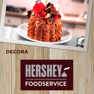 Decora.  Sobre un fondo con textura de madera se ve una fotografía de un postre de Chocolate decorado con frutos rojos, una rosa y chocolate.